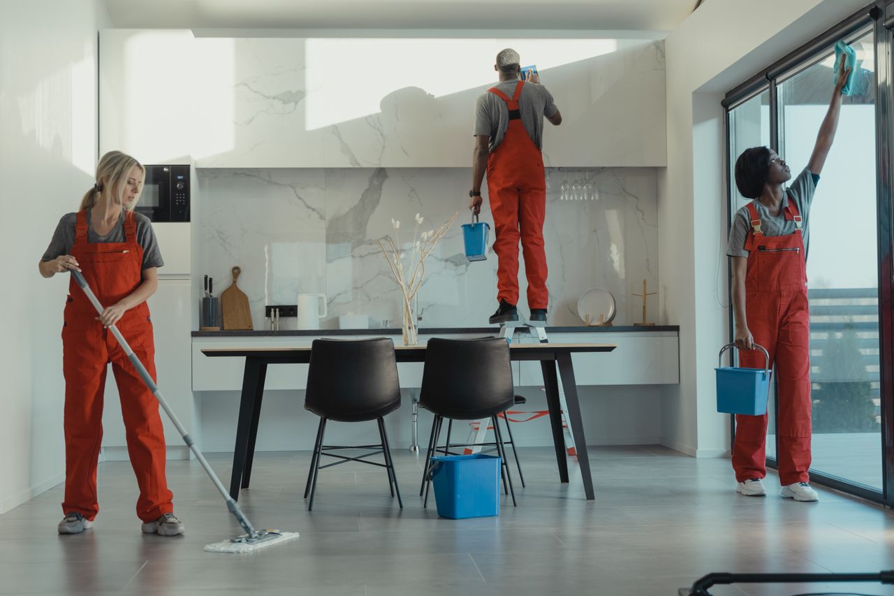Groupe de nettoyeurs en uniformes rouges en train de passer la serpillière et d’essuyer les vitres dans une salle à manger moderne.