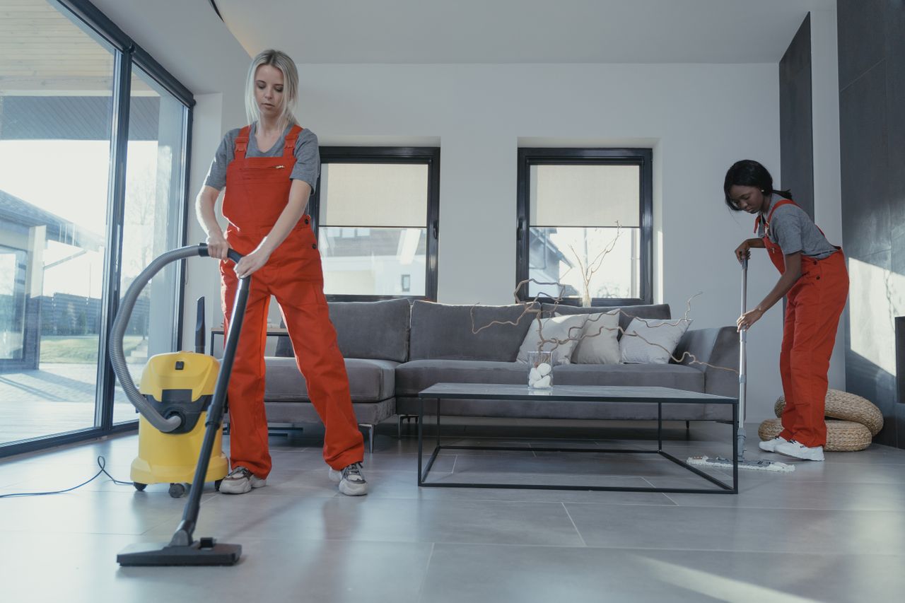 services-01 Two women in red overalls vacuuming and sweeping a modern living room.